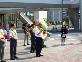 駅前の横断歩道の前の広場にマスコットキャラクターかいちゃんと黄色のタスキを肩にかけた関係者が立ち、中央に右手にマイクを持って話をしている市長の写真