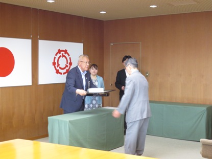 国旗と三郷市旗が飾られた室内で、スーツを着た上野さんと市長が向かい合って立ち感謝状の贈呈を行っている様子の写真