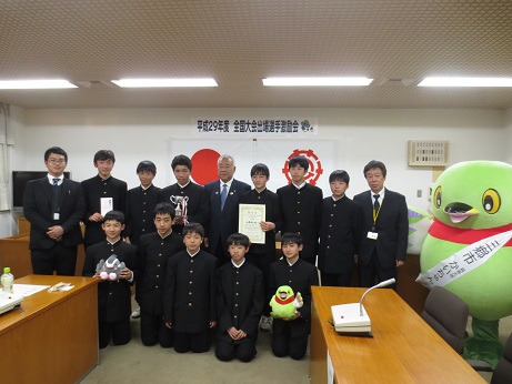 「平成29年度全国大会出場選手激励会」のポスターと国旗・三郷市旗が掛けられている壁の前で、それぞれかいちゃん・つぶちゃんのぬいぐるみ、トロフィー、賞状を持った男子生徒を含める男子チームのメンバーと引率の先生が、三郷市長とかいちゃんとともに並んで記念撮影をしている写真
