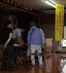 「災害用伝言ダイヤル171」と書かれた、のぼりが設置されたコーナーで担当の男性に説明を受けている2名の女性の写真