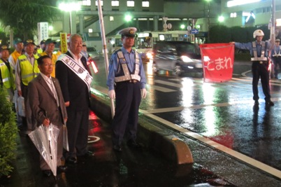 日が暮れた雨天のなか、道路上で「とまれ」と書かれた赤旗を持っている警察官が検問を行っている傍で、市長と吉川警察署長など数名が歩道上に立ち通行する車を見ている様子の写真