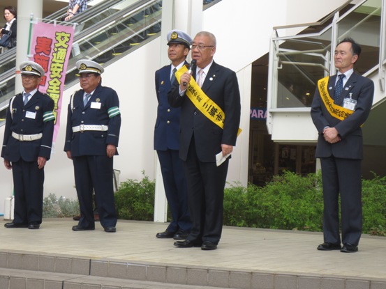商業施設の屋外の会場で、黄色のタスキを身に着けた市長がマイクを手に挨拶をしている写真