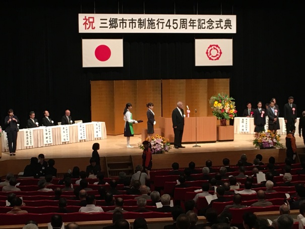 「祝三郷市市制施行45周年記念式典」と書かれたボードが国旗と三郷市旗とともに吊るしてあり、色とりどりの花々が飾られたステージの演台の近くに三郷市長と女性2名が並び、ステージ上に配置された席に座る来賓者の前には受賞者の人達が並んでいる舞台の写真
