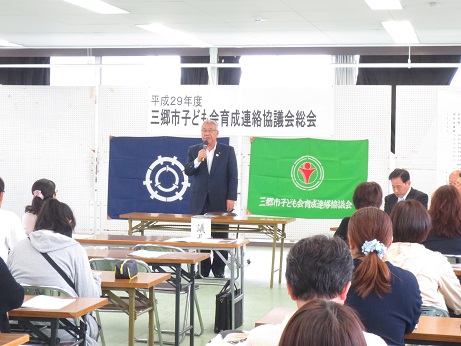 「 平成29年度三郷市子ども会育成連絡協議会総会」と書かれたポスターと紺色の三郷市旗、緑色の三郷市子ども会育成連絡協議会総会の旗が貼りつけられた白いボードの前で議長としてあいさつの言葉を述べる三郷市長と、席に座り話を聞く参加者の写真