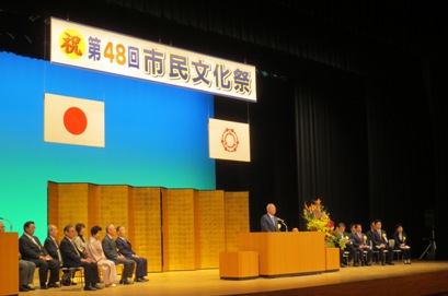 「祝 第48回市民文化祭」と書かれた横断幕や国旗と三郷市旗、金屏風が立てかけられたステージの檀上で挨拶を行っている市長の写真