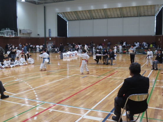 体育館の一画で、四方から関係者や選手たちが見守る中、2名の子どもが試合をしている様子の写真