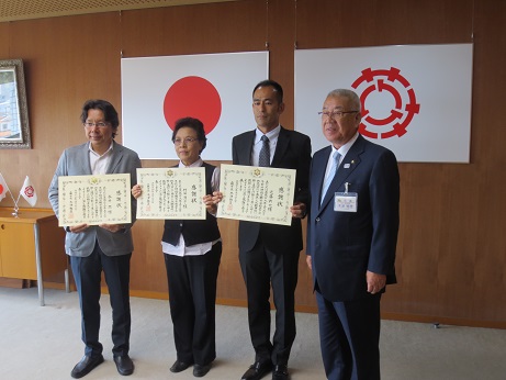 国旗と三郷市旗が吊るされた壁の前で、竹田清子さん、近藤新也さん、永井潤さんが感謝状を手に持ち三郷市長とともに記念撮影をしている写真