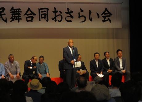 「民謡・民舞合同おさらい会」と書かれた横断幕が吊るされているステージ上で、三郷市長が資料を手に持ちながら立って話をし、ステージ上に配置された赤い長椅子に座る関係者と観客席に座るたくさんの参加者が話を聞いている写真