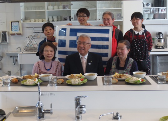 ギリシャ料理が並べられたテーブルのそばで、三郷市長がギリシャ国旗を手に持った関係者とともに記念撮影をしている写真