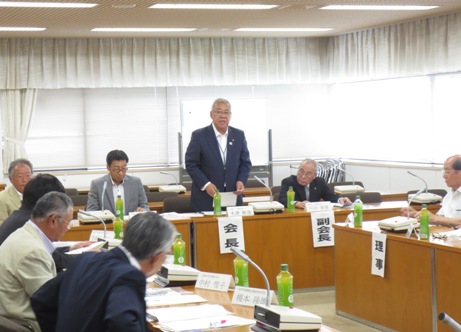 総会での役割が書かれた紙が各席の側面に貼られ、机の上には席札とお茶のペットボトルが置いてあり、「会長」と書かれた席を立ちあいさつの言葉を述べる三郷市長と、席に座り手元の資料に目を通しながら話を聞く総会メンバーの写真