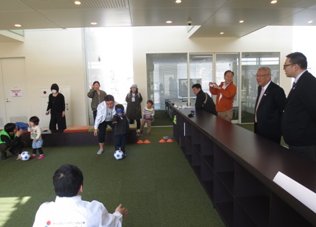 サッカーボールのそばで右手を前に伸ばして立ちブラインドサッカーを体験する青いアイマスクを付けた男の子と、その様子を見守る三郷市長と関係者の様子