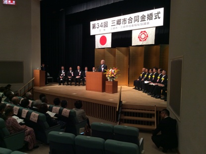 「第34回三郷市合同金婚式」と書かれた横断幕の下のステージの檀上に市長が立ち挨拶を行っている写真