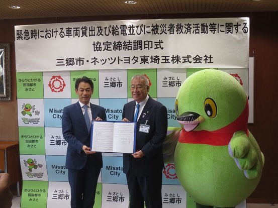 「緊急時における車両貸出及び給電並びに被災者救済活動等に関する協定締結調印式」と書かれたポスターが貼りつけられている三郷市のボードの前で、ネッツトヨタ東埼玉株式会社の男性と三郷市長が協定書をともに持ち、かいちゃんと一緒に記念撮影をしている写真