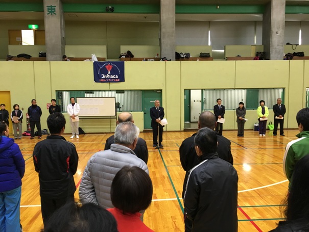 体育館の中でマイクの前に立ち、正面を向いて挨拶をする市長と、その後方両側に一列に並んで立つ関係者、市長の方を向いて話を聞く参加者たちの写真
