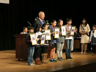 ステージの中央に市長と受賞した6名の子どもたちが作品を持って横一列に並んで立っている写真