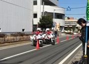 道路の中央線に赤いコーンが並べられ、コーンの間を2台横に並んだ交通機動隊の白いバイクが走行している様子の写真
