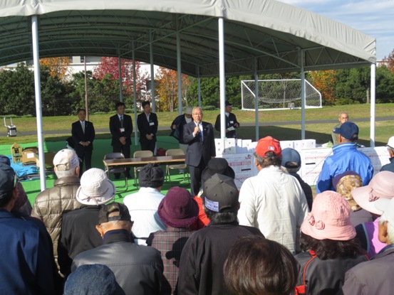 公園の屋根付き広場前に配置され景品が置かれた長机の前に立ち、挨拶の言葉を述べる三郷市長と、市長と向かい合って立ち話を聞く大勢の参加者の写真