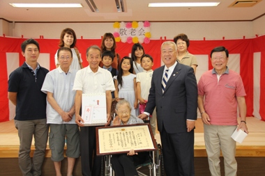 敬老会と書かれ紅白幕が貼られた室内で、車いすに座って額縁に入った賞状を持った笑顔の鈴木まつさんと市長、7名の大人と4名の子どもの家族が並んで立っている記念写真