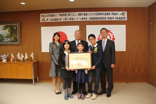 「平成26年度優れた「早寝早起き朝ごはん」運動の推進にかかる文部科学大臣表彰」と書かれた横幕の下に国旗と三郷市市旗が飾られた室内で、額に入った表彰状を持っている受賞した3名の児童と幡谷先生、青木校長先生の5名と市長が並んで立っている写真
