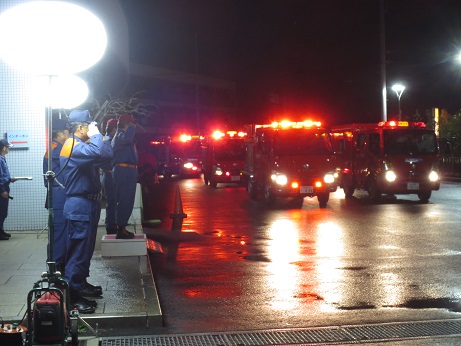 赤色灯を灯したたくさんの消防車が、2列に並んで夜の特別警戒に出発する様子と、それを手前で敬礼しながら見送る制服を着た消防団員たちの写真