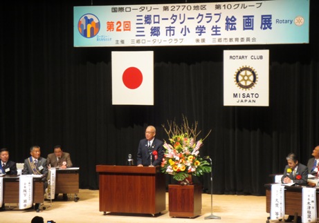 「第2回三郷ロータリークラブ三郷市小学生絵画展」と書かれたボードと国旗・三郷ロータリークラブの旗が吊るされているステージ上で、赤・ピンク・オレンジ・黄色・白の豪華な花々で彩られた花台と並ぶ演台に立ち挨拶の言葉を述べる三郷市長と、ステージの両側に配置された席に座り話を聞く胸章を付けた来賓者の写真