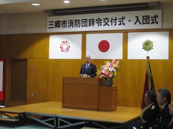 「三郷市消防団辞令交付式・入団式」の吊り看板が設置されている舞台上で、演壇に立って挨拶をしている市長の写真