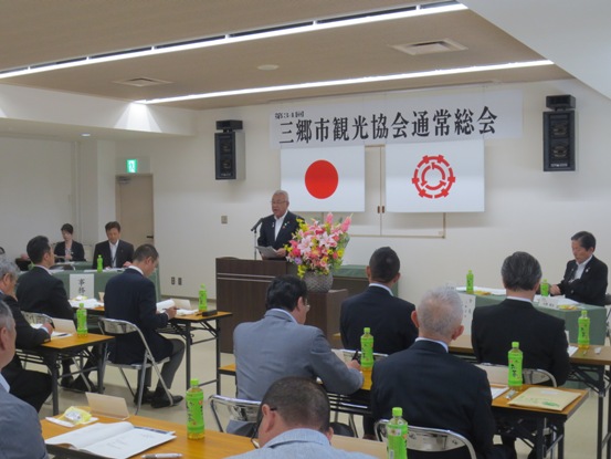 壁に「第34回三郷市観光協会通常総会」の紙が貼られた部屋で、等間隔に並べられた長机に2名ずつの関係者が椅子に着席しているなか、演壇で挨拶をしている市長の写真