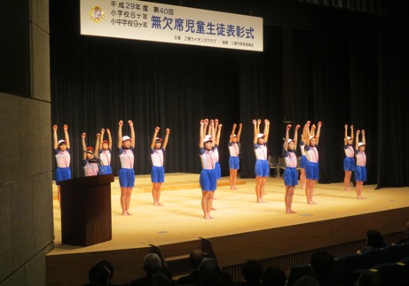 「平成29年度第40回小学校6ヶ年、小・中学校9ヶ年無欠席児童生徒表彰式」と書かれた横断幕が吊り下げられているステージ上で、紅白帽と体操服を着用した子どもたちが等間隔に並び両手を真上に挙げラジオ体操を披露している写真