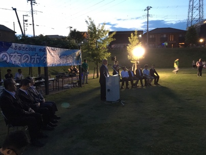 日が暮れて奥に立っている樹木の前に大きな照明が照らされた会場で、「夏の夜のホタル観賞会」と書かれた横断幕の前に設置された演壇で挨拶を行っている市長の写真