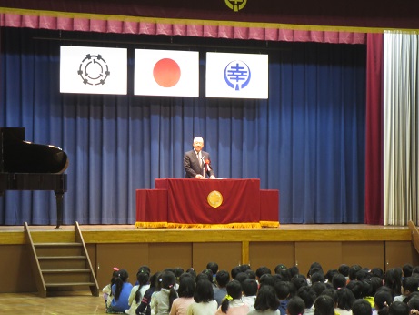 赤色の装飾カバーが施されたステージ上の演台に立ちお祝いの挨拶の言葉を述べる三郷市長と座って話を聞く子どもたちの写真
