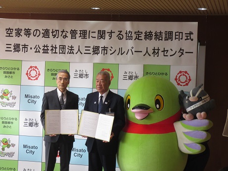 「空家等の適切な管理に関する協定締結調印式」と書かれたポスターが貼りつけられている三郷市のバックボードの前で、三郷市長と 公益社団法人三郷市シルバー人材センターの男性が協定書を手に持ち、かいちゃん・つぶちゃんとともに記念撮影をしている写真