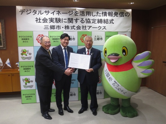 三郷市のバックボードに「デジタルサイネージを活用した情報発信の社会実験に関する協定締結式」と書かれた紙が貼られている部屋で、阿部様や菊地社長と一緒に協定書を持っている市長と、横にかいちゃんが並んでいる写真