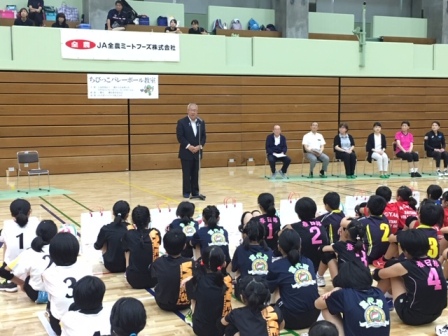 「ちびっこバレーボール教室」と書かれた横幕が壁に貼られた体育館で、右側に6名の関係者が座り、手前に体育座りしたユニホームを着た子供たち、中央に設置されたマイクスタンドの前で挨拶を行っている市長の写真