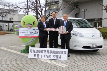 「電気自動車活用事例創発事業日産e-NV200引渡式」と書かれた看板と白色の車の間に市長と2名の男性が一緒に大きなゴールドキーを持って立ち、左側のかいちゃんと一緒に並んでいる写真