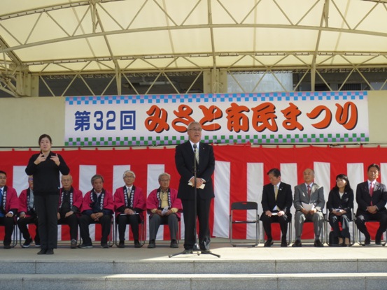「第32回三郷市民まつり」と書かれた横断幕の下に紅白幕が掛けてあるステージ上で、挨拶の言葉を述べる三郷市長と近くに立って手話通訳をする女性、ステージ後方に並べられたパイプ椅子に座り話を聞く関係者の写真