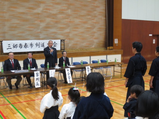 「三郷市春季剣道大会」の横断幕が設置されている体育館で、剣道着を着た剣士のかたたちが整列して正面の市長の挨拶を聞いている写真