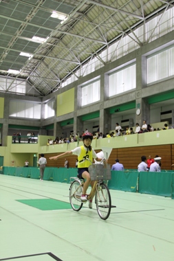赤色のヘルメットを被っている児童が自転車に乗って右手を横に真っ直ぐ伸ばしている大会の様子の写真