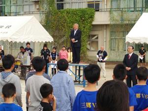 左右にテントが設置された前谷小学校の校庭の朝礼台に立ち挨拶を行っている市長の話を聞いている子供たちの後ろ姿の写真