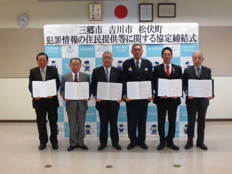 「三郷市 吉川市 松伏町 犯罪情報の住民提供等に関する協定締結式」と書かれたバックパネルの前で、3名の市長と町長、警察署の代表他2名の関係者の男性が協定書を持ち横一列に並んでいる記念写真