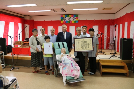 車いすに座っている山田みよさんと7名の家族と市長がステージの前に並んでいる記念写真