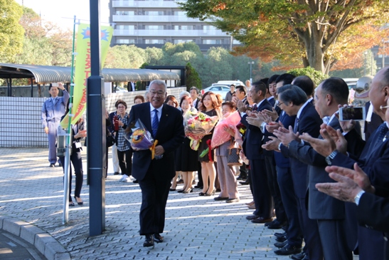 紫色のラッピングペーパーで包まれたオレンジ色の花束を持ち笑顔で歩く三郷市長と、歩道の端に並んで立ち拍手をして市長を出迎える多くの市民、議員、職員の写真