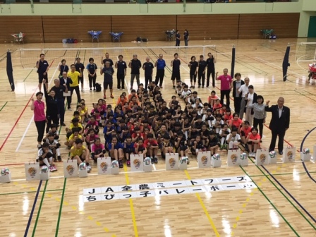 体育館の床に「JA全農ミートフーズちびっこバレーボール」と書かれた文字が貼られ、奥にチームごとに体育座りしている子供たち、周囲にガッツポーズを行っている関係者や市長が立っている写真