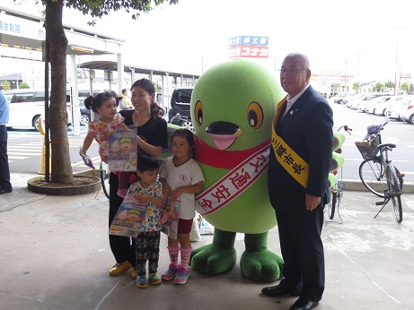 商業施設前の通路で市長とマスコットキャラクターかいちゃん、3名の子どもと母親が一緒に並んでいる写真