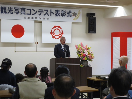 「観光写真コンテスト表彰式」と書かれた横断幕と国旗・三郷市旗が吊るしてある壁の前で、ピンク・黄色・白の豪華な花々で彩られた花台と並ぶ演台に立ち挨拶の言葉を述べる三郷市長と、席に座り話を聞く受賞者の写真