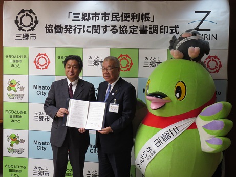 「三郷市市民便利帳」協働発行に関する協定調印式と書かれた三郷市のパネルの前で、調印した協定書を正面に向けて一緒に持つ市長と株式会社ゼンリンの代表者、市長の横に立つかいちゃんとつぶちゃんの記念写真