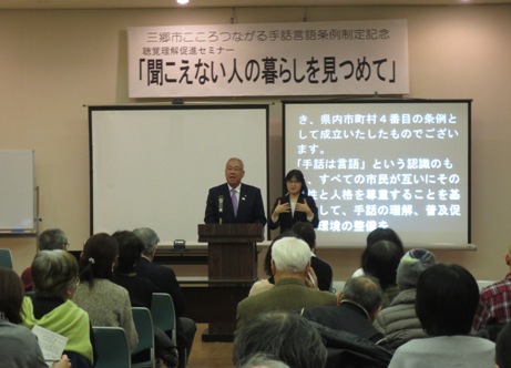 壁に「聞こえない人の暮らしを見つめて」と書かれた看板が掲げられており、その下にある2枚のスクリーンの向かって右側に文章が映し出されている様子と、その前に置かれた演台に立ち、たくさんの参加者の方を向いてあいさつをする市長、隣で手話通訳をする女性を写した写真