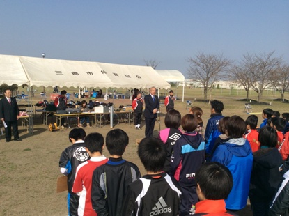 テントの前に設置された小さな台の上に立っている市長の話を向かい合って立っている子供たちが聞いている後ろ姿の写真