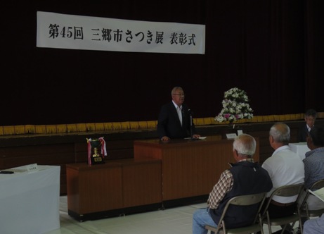 「第45回三郷市さつき展表彰式」と書かれた横断幕がステージ上の舞台幕に貼り付けられてあり、置台にはトロフィーが飾られ、さつきの花が置かれた演台に立ち話をする三郷市長とパイプ椅子に座って話を聞く参加者の写真