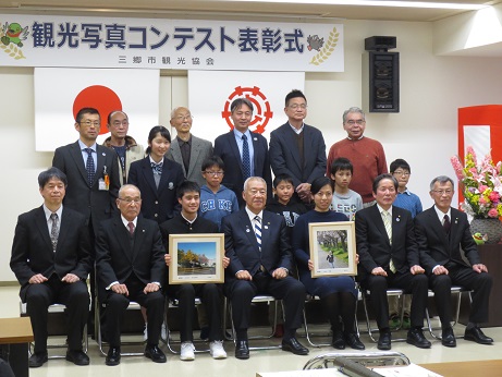 「観光写真コンテスト表彰式」と書かれた横断幕と国旗・三郷市旗が吊るしてある壁の前で、三郷市長が自分の作品を手に持った女性と男子学生、その他の受賞者とともに並んで記念撮影をしている写真