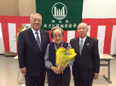 「埼玉県みさと団地自治会」と書かれた旗と紅白幕が貼られた前で海瀬会長と市長が花束を持った碓井あや子さんと並んで立っている記念写真